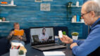 An older man speaks with his doctor over the computer while his wife reads in the background