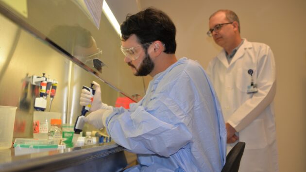 A scientist studying hepatitis B virus (HBV) works under the hood in a lab as another scientist in a white coat looks on.