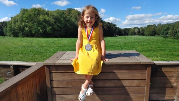 young girl in a yellow dress with a champion medal around her neck, sitting on a wooden platform on a golf course