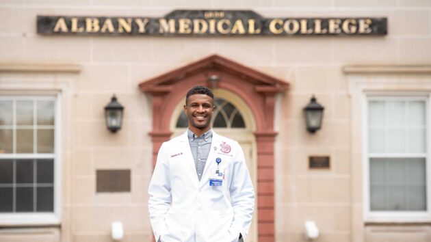 medical student in a white coat, standing in front of the Albany Medical College Sign