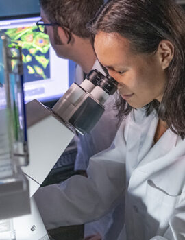 Scientist looking in microscope while colleague looks at cell image on a screen.