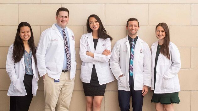 five medical students in white coats leaning up against the building