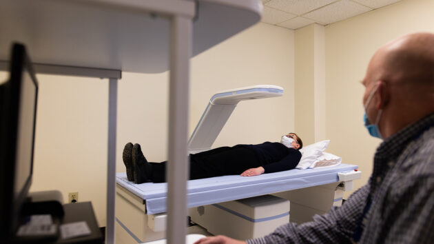 Clinician at controls of scanning apparatus while patient lies on a platform below scanner.