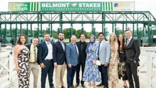 A group of people, dressed formally, stand next to the starting gate at Saratoga Race Course for the Light up the Night event