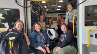 A boy sitting in the back of an ambulance is surrounded by his care team. One woman is holding a graduation cap and gown.