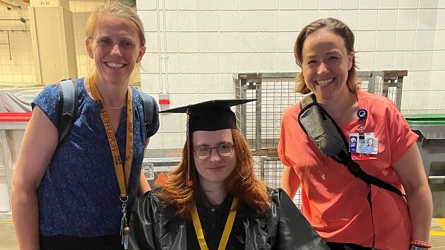 A doctor and nurse stand on either side of a boy in a wheelchair. The boy is wearing a graduation cap and gown