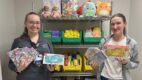 Two child life specialists holding Nintendo Switch consoles and games, standing in front of a shelving unit filled with toys and books