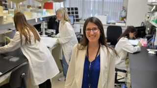 Research scientist Kristen Zuloaga, PhD stands in her lab while three people work behind her