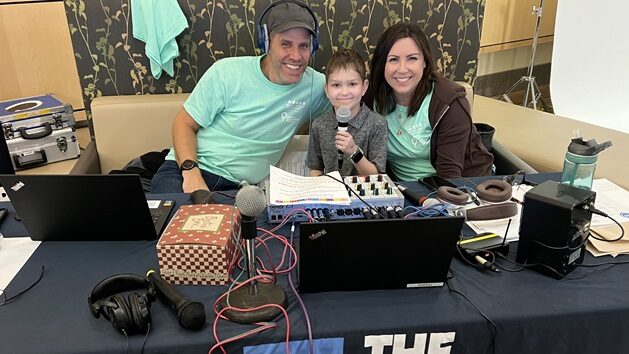 A young boy holding a microphone, sitting with two radio hosts