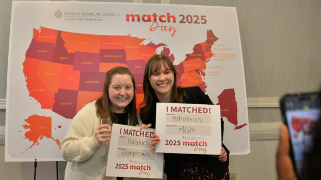 Two medical students stand in front of a map of the U.S. on Match Day 2025, holding signs showing where they matched